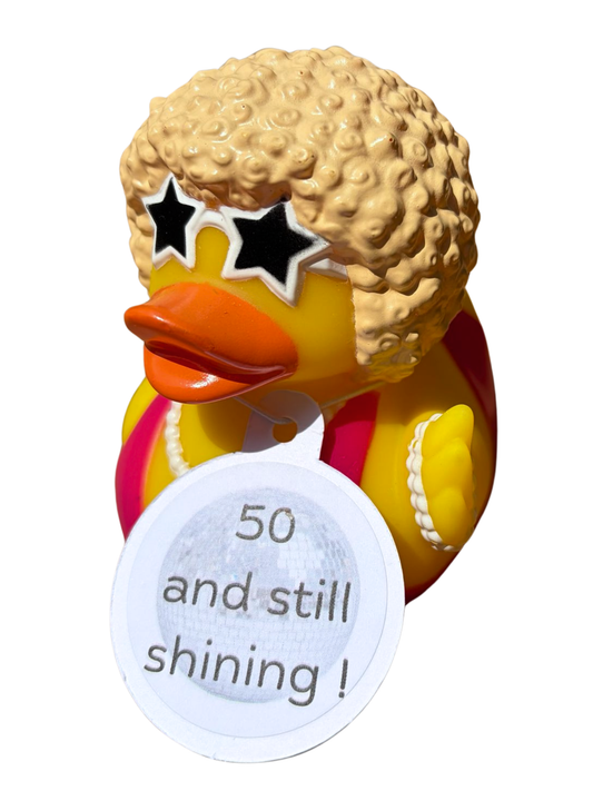 Gele badeend met blond krulhaar, stervormige zonnebril en een hangtag met de tekst ‘50 and still shining!’.