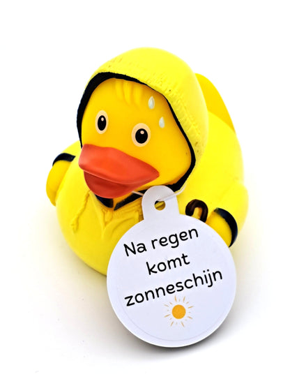 Gele badeend in een gele regenjas met capuchon en zwarte randen, met een witte hangtag met een zonnetje en de tekst ‘Na regen komt zonneschijn’.