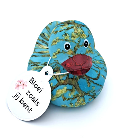 Blauwe badeend met Van Goghs Amandelbloesem als patroon, donkerrode snavel en een witte hangtag met de tekst ‘Bloei zoals jij bent’.